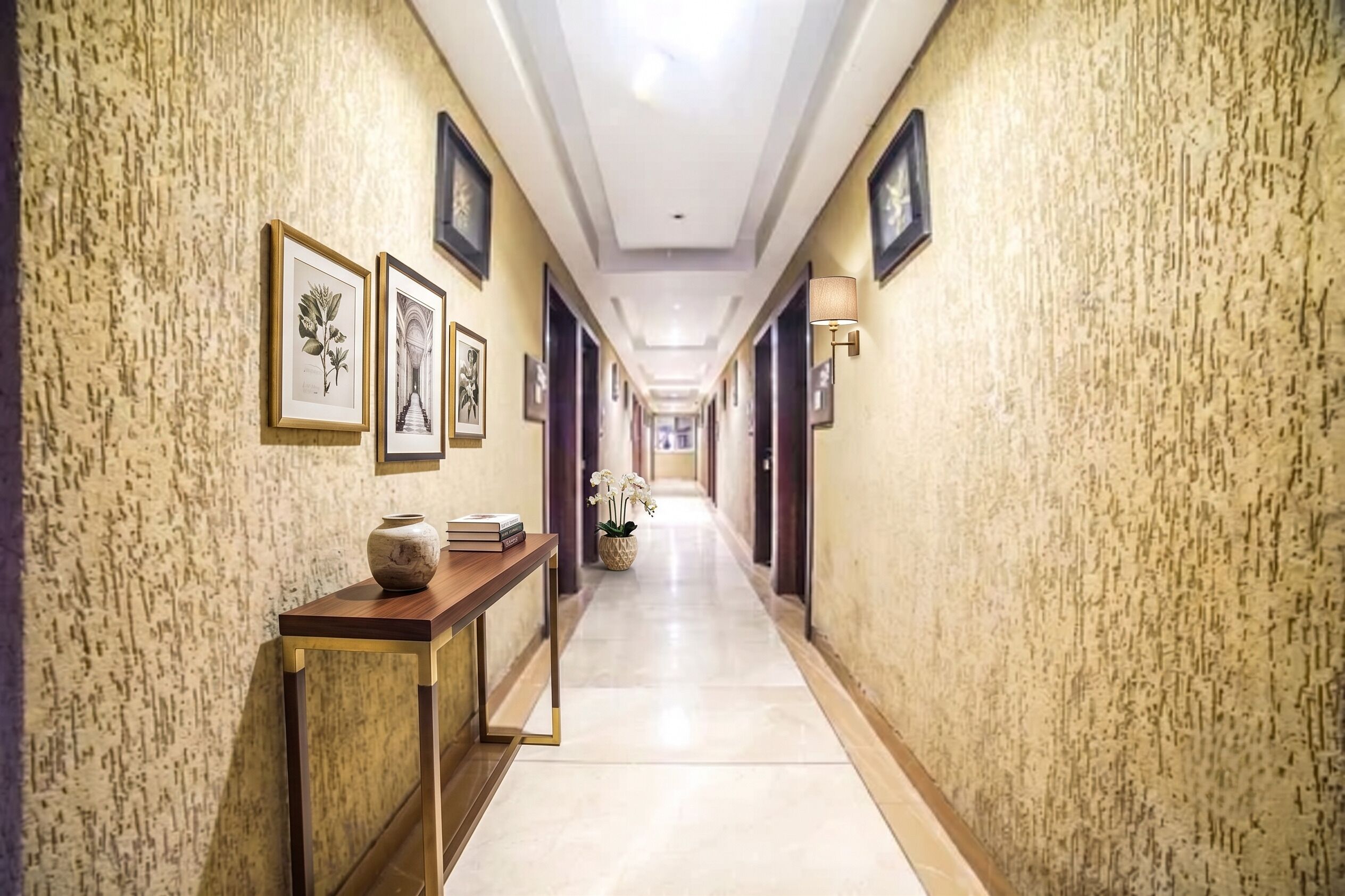 Un pasillo de hotel con impresiones botánicas enmarcadas en las paredes, una consola de madera con un florero y libros, y una orquídea en maceta.