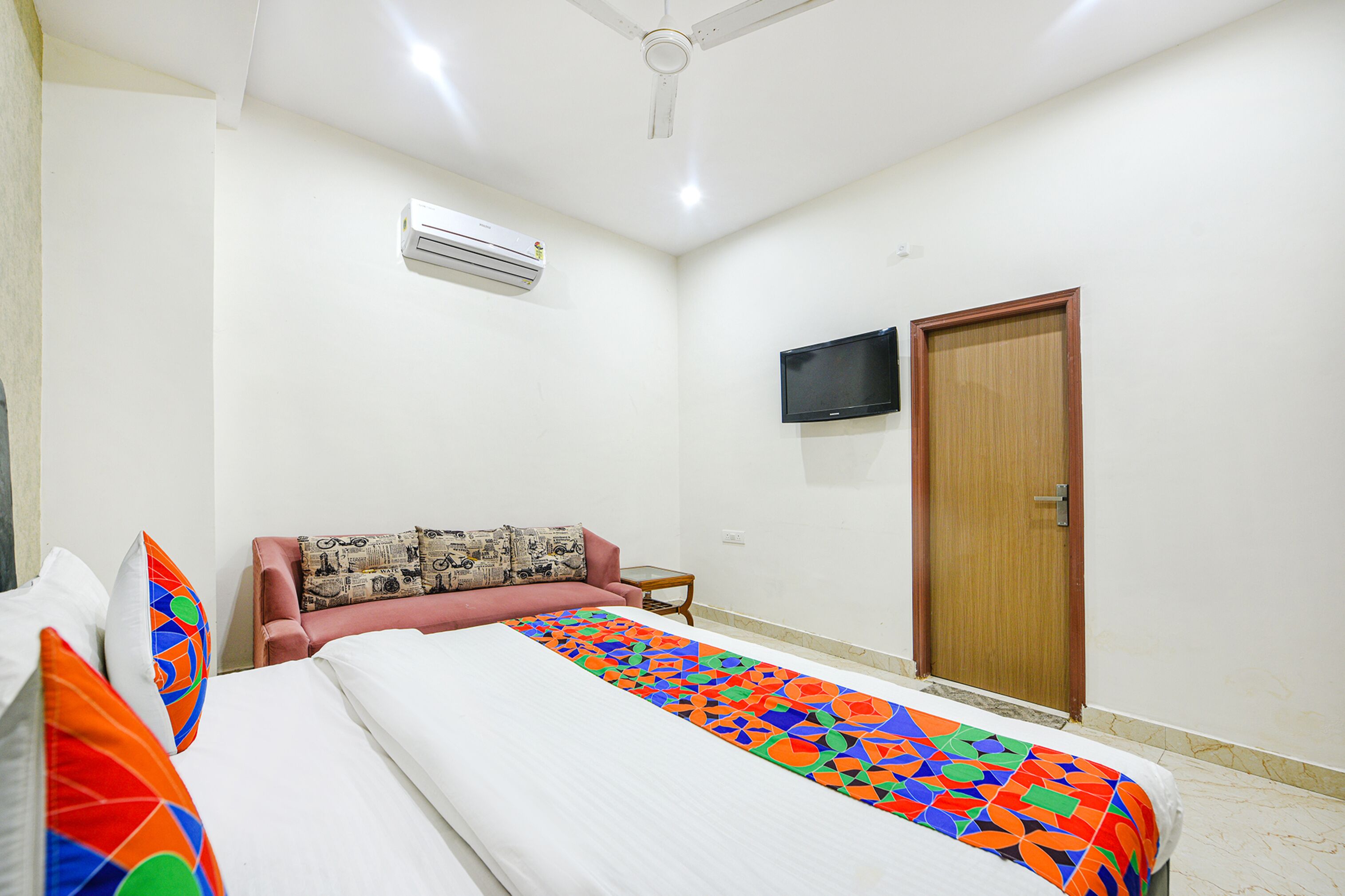 A hotel room with a bed, a television mounted on the wall, an air conditioning unit, and a wooden door.