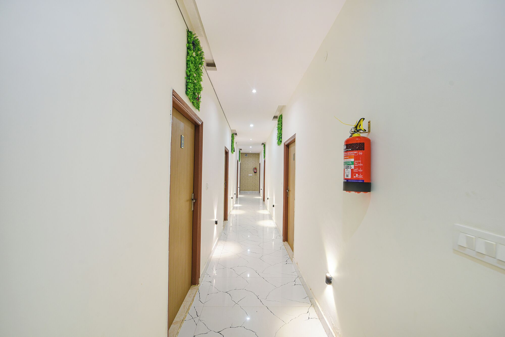 Un couloir aux murs blancs, avec des portes en bois et un extincteur fixé au mur.