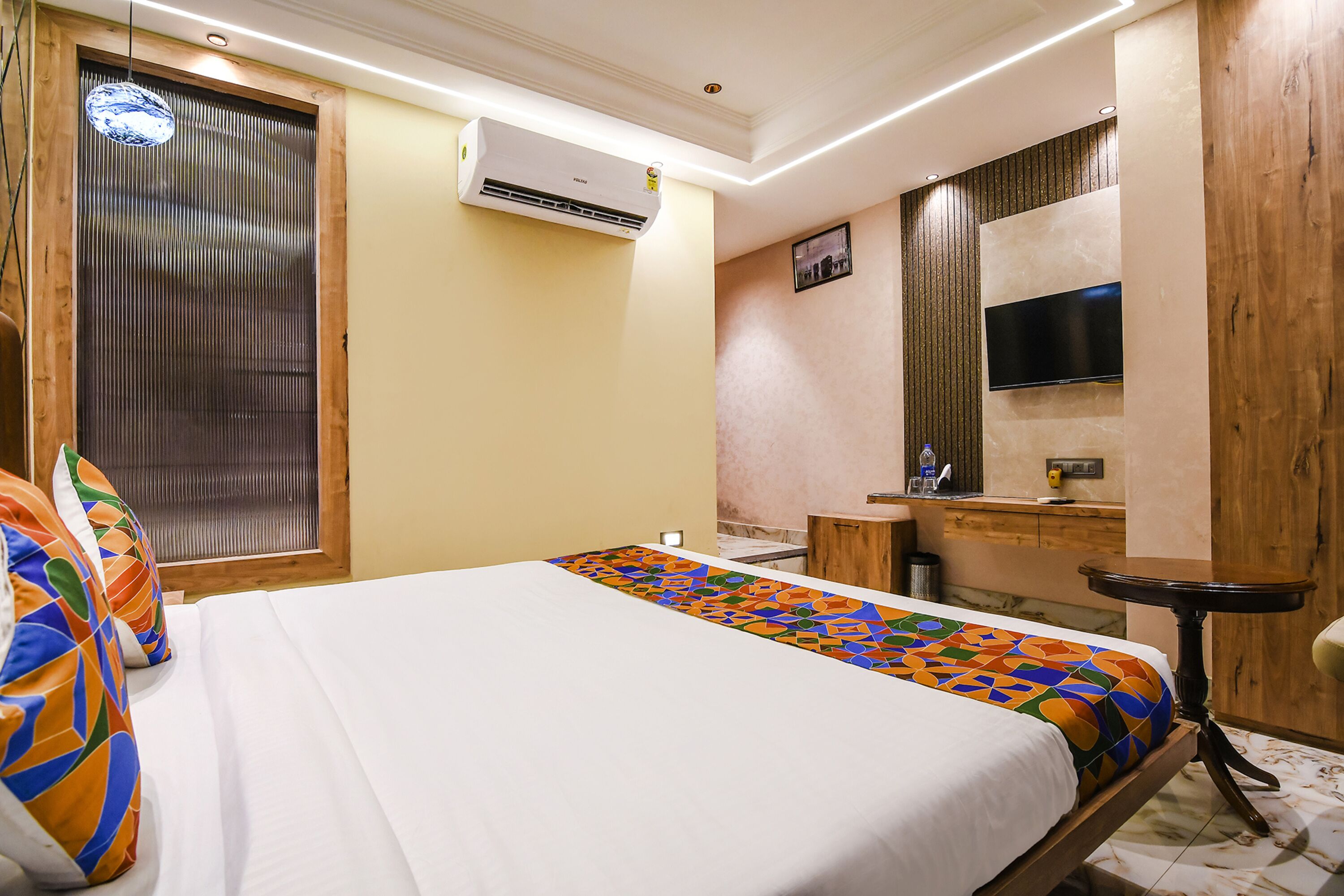 Quarto de hotel com cama, cabeceira de madeira, ar-condicionado, televisão fixada na parede e uma pequena mesa redonda.