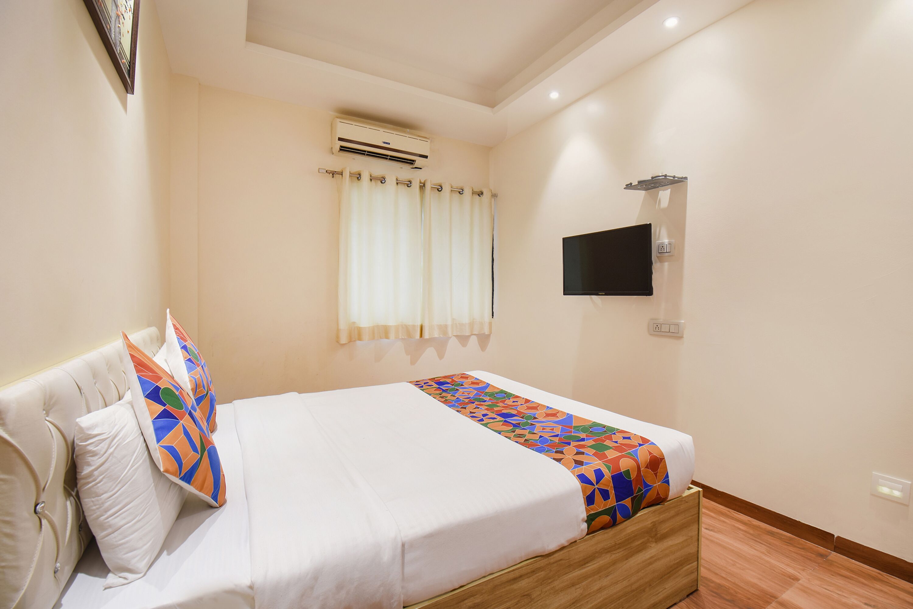 A neatly made bed with a colorful blanket, a flat-screen TV mounted on the wall, and an air conditioning unit above the window.