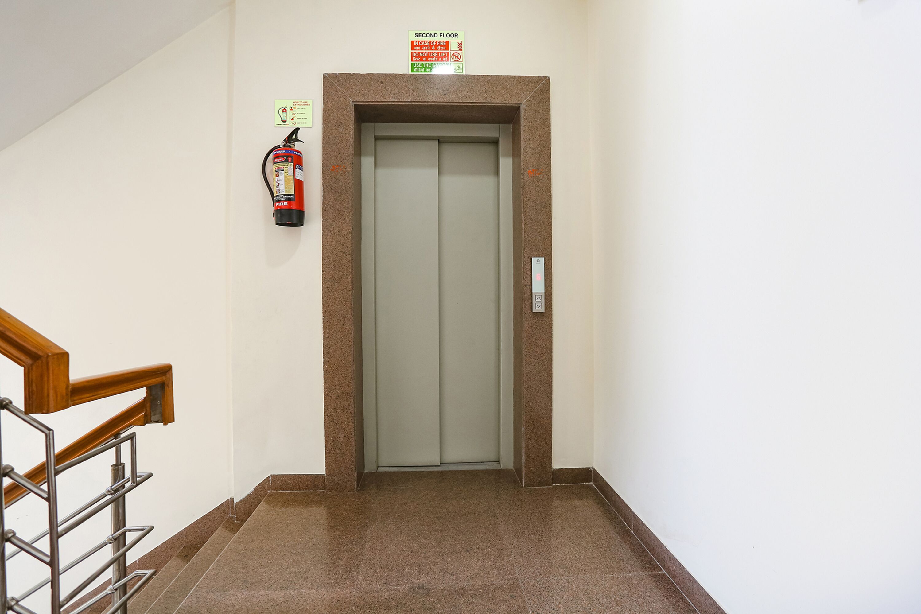 Un ascenseur avec une pancarte indiquant qu’il dessert le deuxième étage, un extincteur fixé au mur et une main courante menant à un escalier.