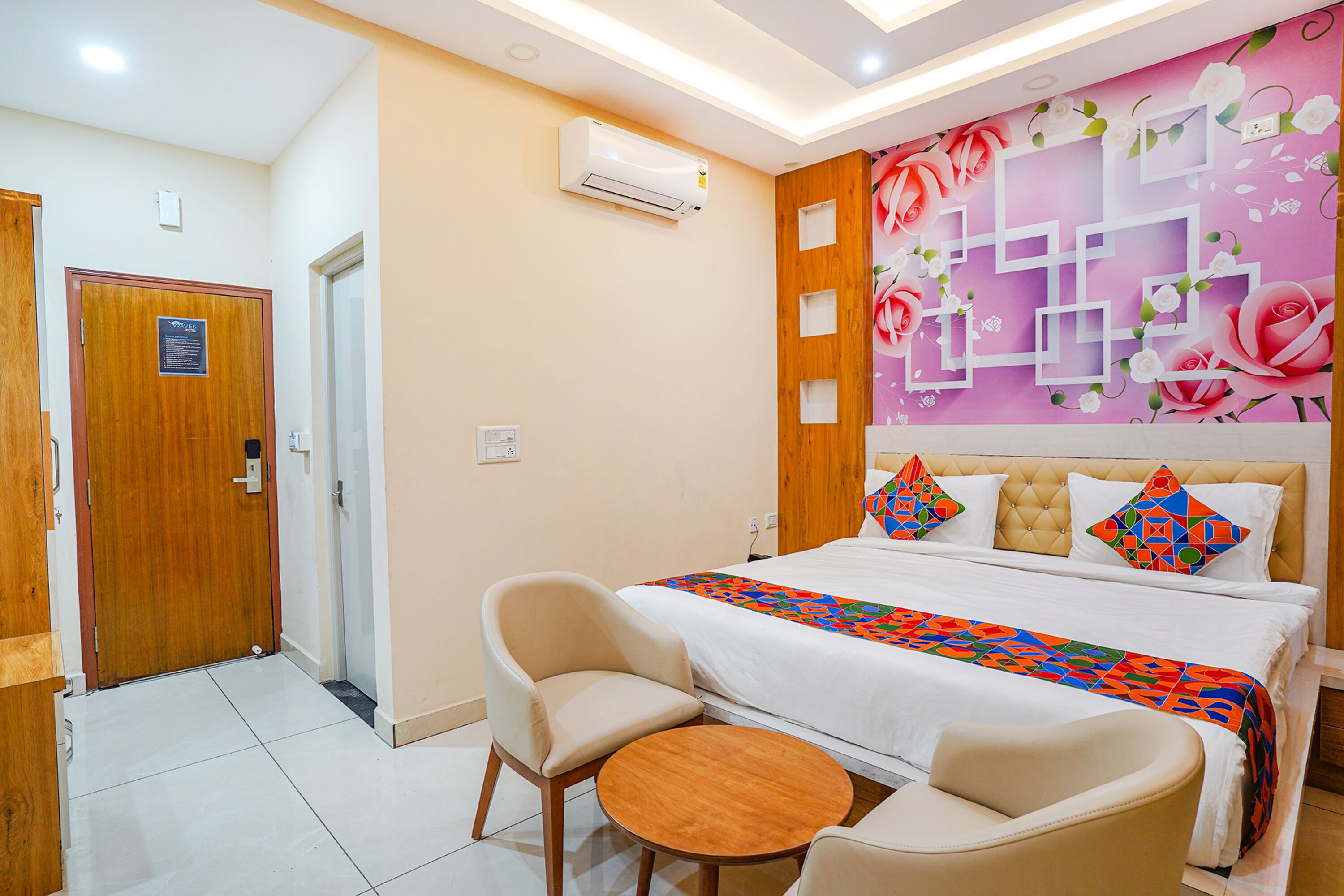 A hotel room with a bed, two chairs, a small table, a wall with floral and geometric patterns, and an air conditioning unit.