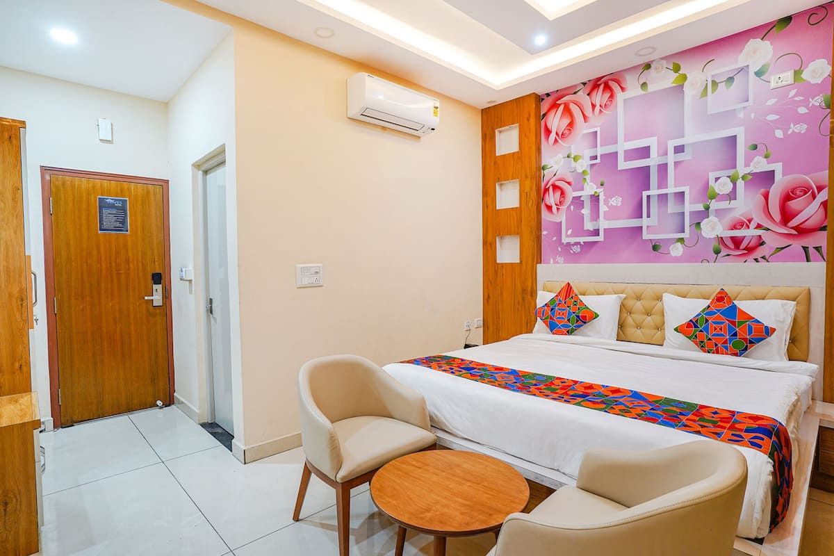 A hotel room with a bed, two chairs, a small table, a wall with floral and geometric patterns, and an air conditioning unit.