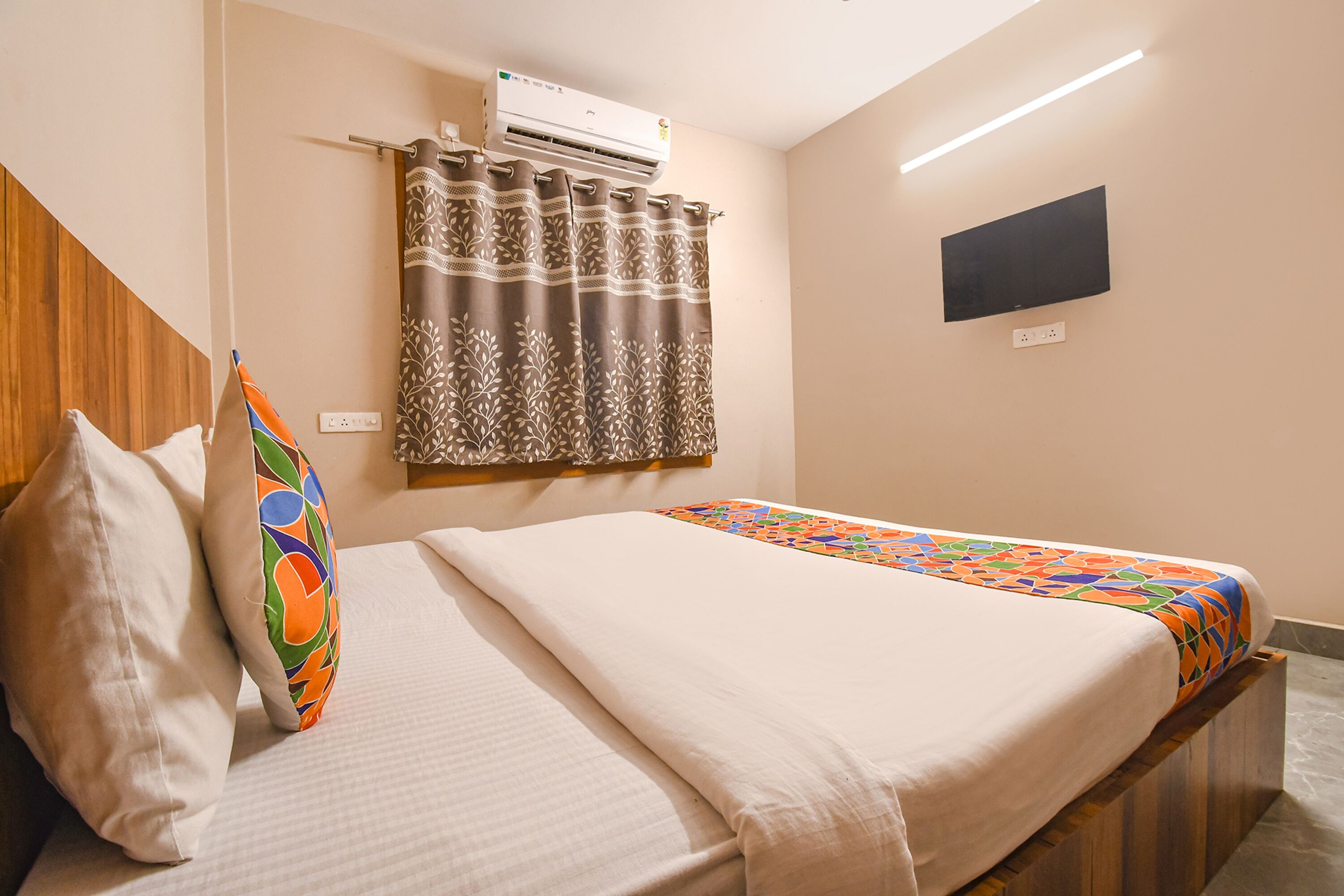 A hotel room with a bed, a pillow, a colorful pillow, a curtain, an air conditioner, a flat-screen TV, and a wall socket.