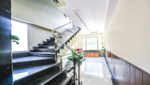 Un'accogliente hall di un hotel moderno, con una scalinata che conduce ai piani superiori, una reception e un'ampia vetrata che lascia entrare la luce naturale.