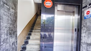 Un ascenseur moderne aux finitions métalliques, un escalier dont les marches alternent entre le noir et le beige, et un panneau indiquant l’obligation de porter un masque.