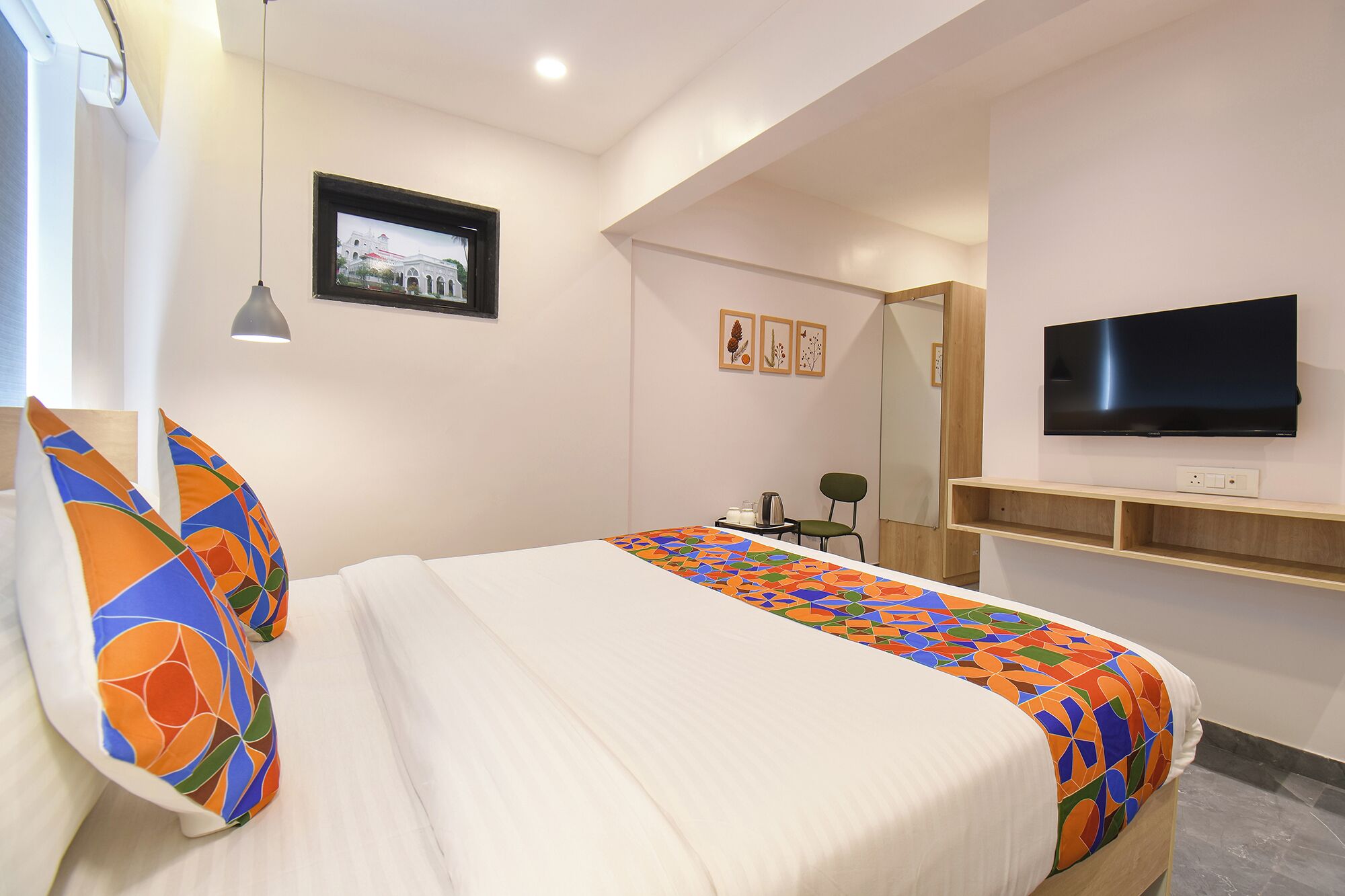A hotel room with a bed, a television, a small table with a chair, and a wall-mounted shelf.