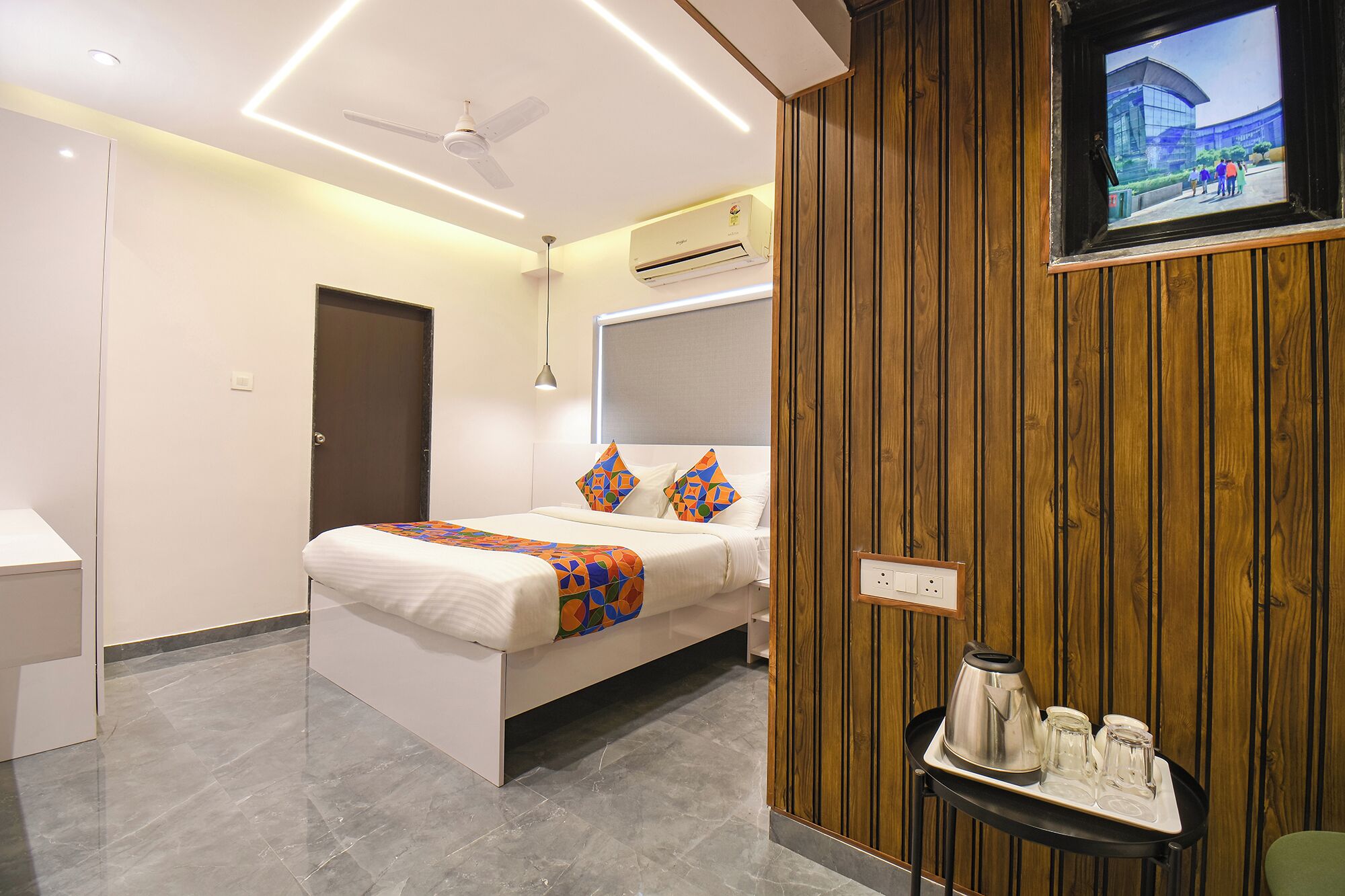A modern hotel room with a bed, a TV mounted on a wooden wall, and a tray with a kettle and glasses.