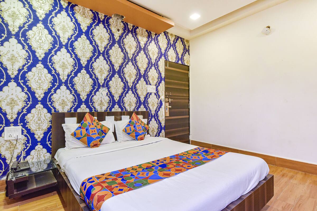 A hotel room with a patterned wallpaper, a bed with white bedding and colorful pillows, a wooden nightstand, and a door with a handle.