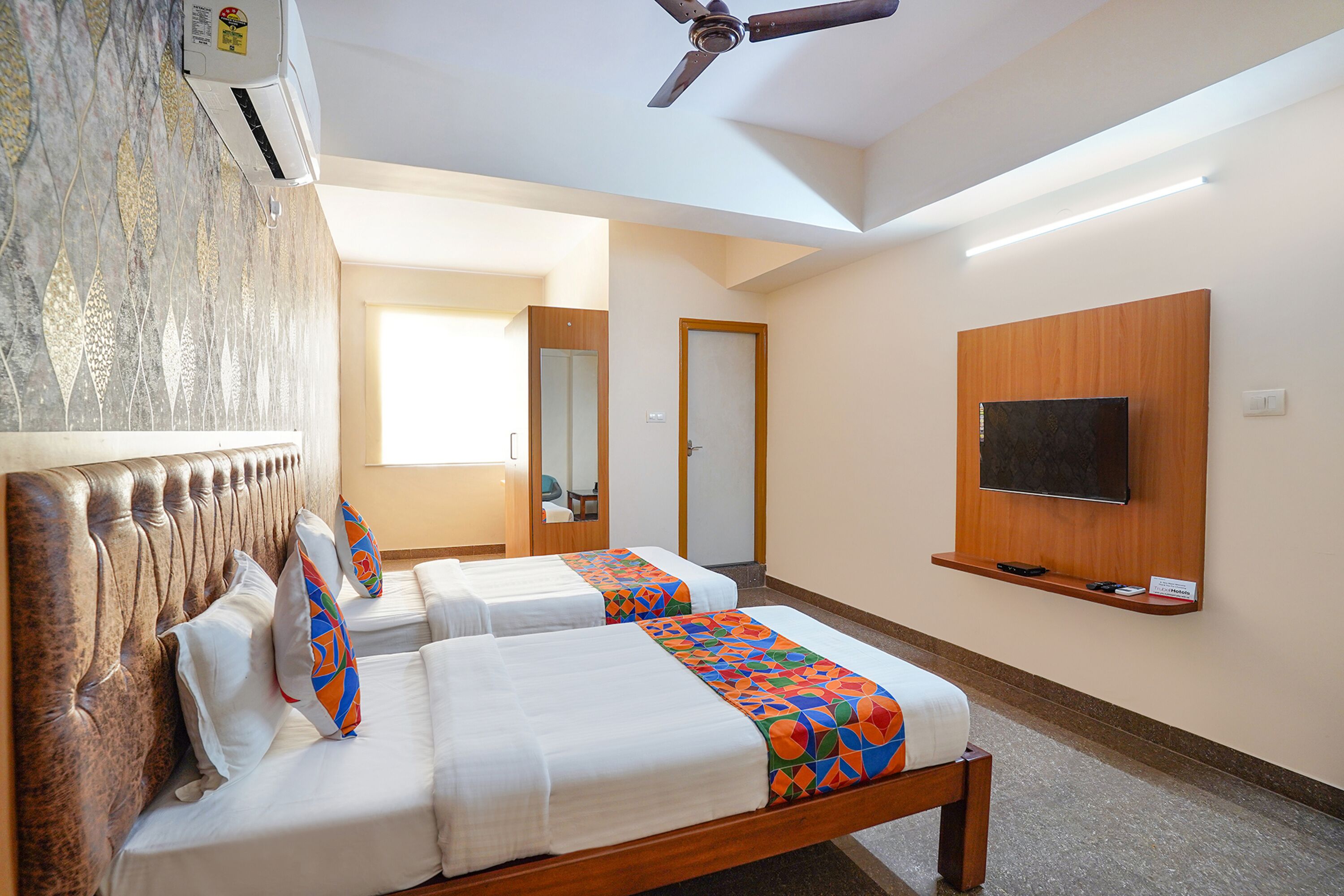 A hotel room with two beds, a ceiling fan, a TV mounted on the wall, and a large mirror.