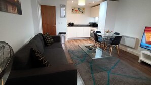 Ein modernes Wohnzimmer mit einer Couch, einem Glastisch und einem Fernseher. Es gibt eine Küchenzeile mit einem Esstisch und Stühlen.