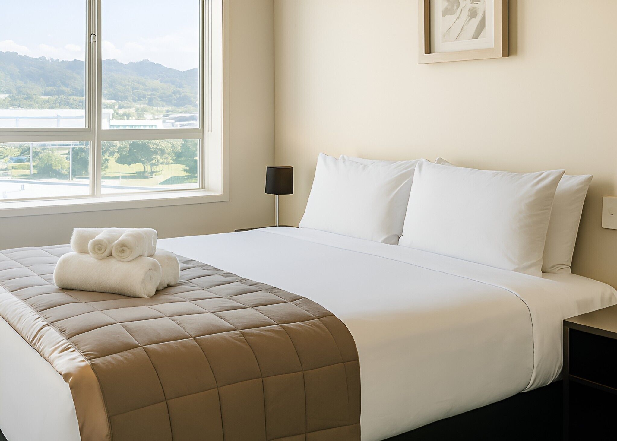 A hotel room with a bed, a window with a view of greenery and mountains, a bedside lamp, and a framed picture on the wall.