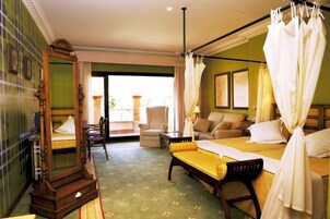 Habitación de hotel con una cama grande, zona de estar con sofá y sillones, una silla de madera, un espejo grande y un balcón con vistas.