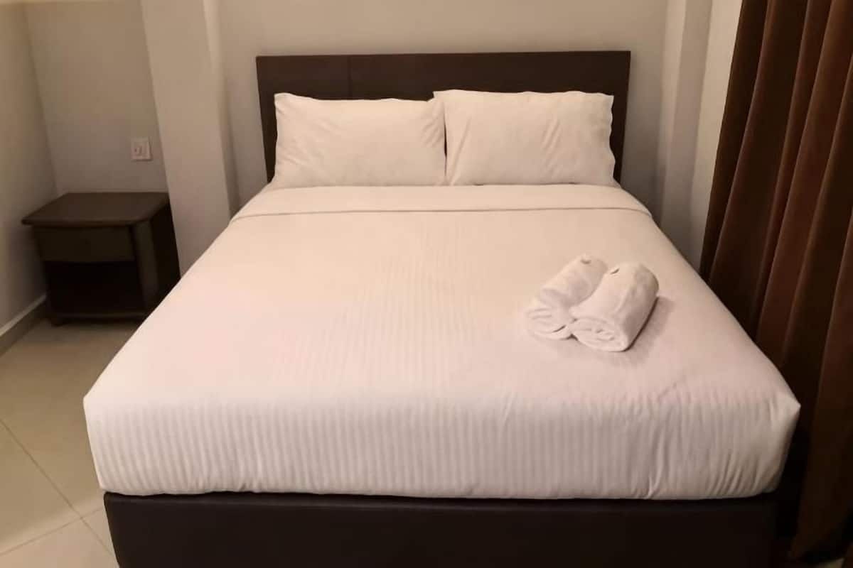 Una cama bien hecha con sábanas blancas y una toalla doblada, un cabecero oscuro y una mesita de noche pequeña.