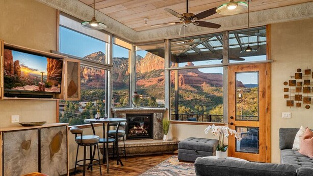 A cozy living room with a fireplace, large windows offering a view of red rock formations, and a ceiling fan. - Signature Uptown Retreat with Epic Views & Hot Tub (Sedona)