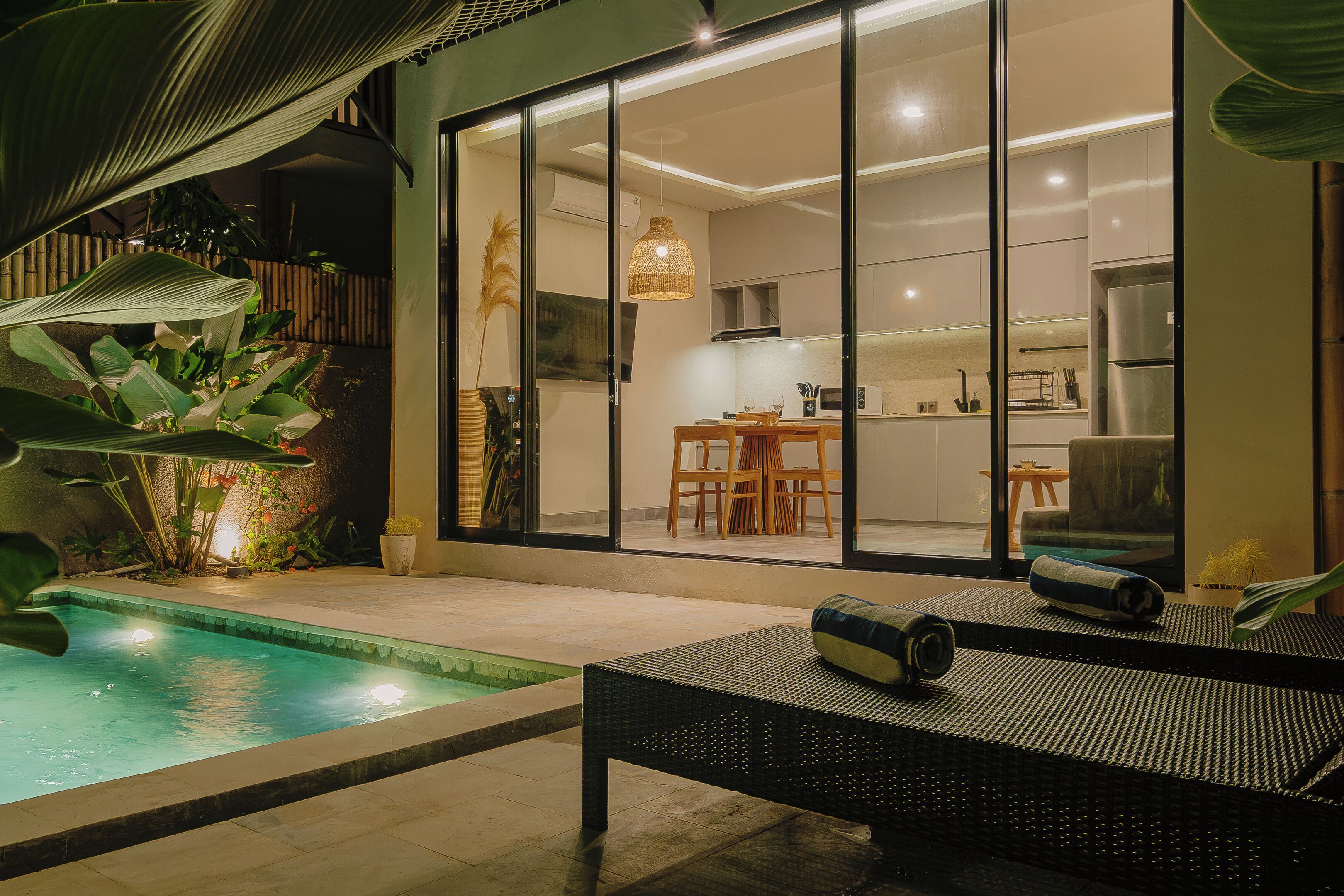 A modern kitchen with a dining area visible through glass doors, a pool with a lounge chair, and a large plant.