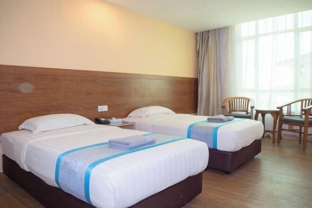 A hotel room with two beds, wooden headboard, a window with curtains, and a small table with chairs.