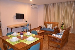 Un salon amĂ©nagĂ© comprenant une table Ă manger pour quatre personnes, un canapĂ©, un tĂ©lĂ©viseur fixĂ© au mur et une fenĂȘtre avec des rideaux.