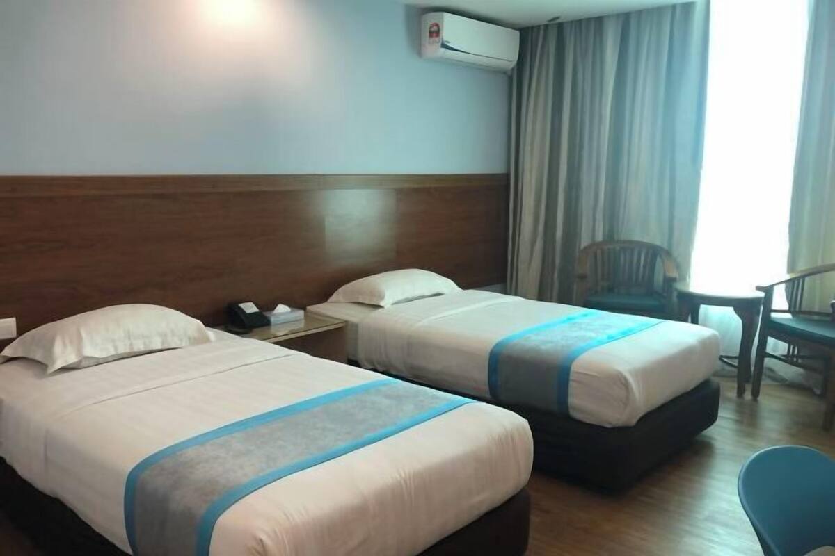 A hotel room with two beds, a desk, a chair, an air conditioning unit, and a window with curtains.