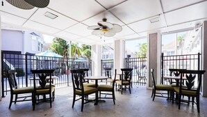 Un área de café al aire libre con mesas y sillas, un ventilador de techo y ventanas amplias.