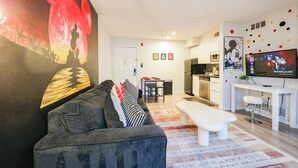 Una sala de estar moderna con un sofá gris, un banco blanco y un tapete colorido. Hay una zona de cocina con una mesa de comedor y sillas, y un televisor mostrando un programa.