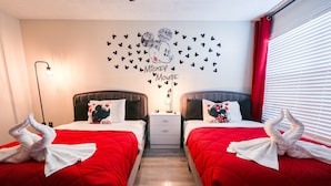 Habitación de hotel con dos camas, ropa de cama roja, decoración de Mickey Mouse en la pared y una mesita de noche con lámpara.