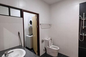 A compact bathroom with a white sink, a toilet, a shower, and a laundry machine visible through an open door.