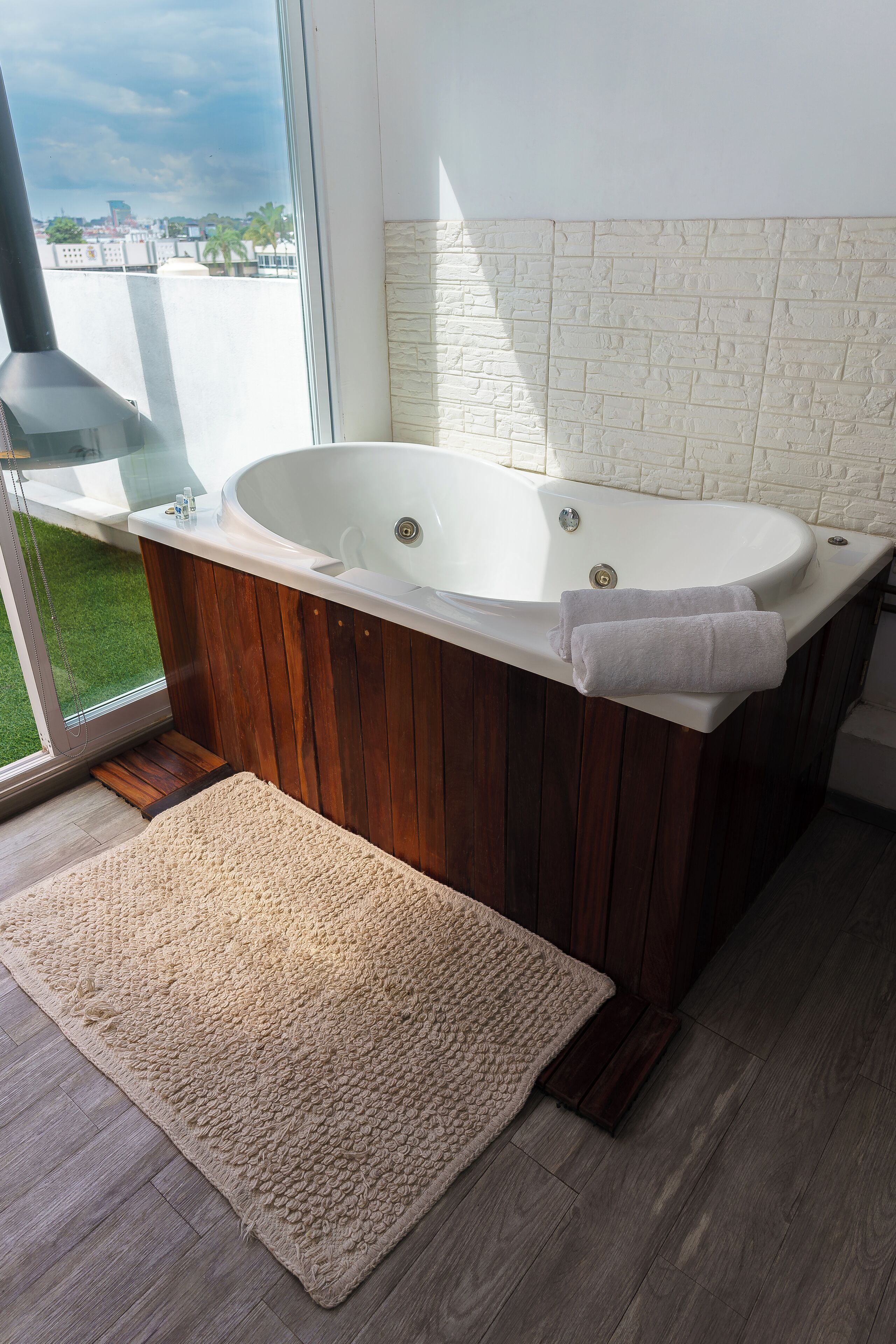 A modern bathroom with a freestanding bathtub, wooden paneling, a large window with a city view, and a beige rug on the floor.