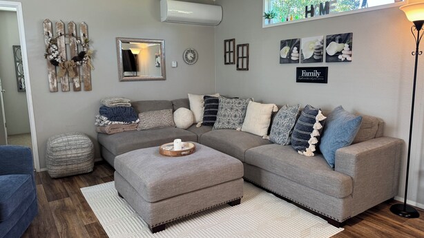 A living room with a grey sectional sofa, a round ottoman, and a white rug. There is a wall-mounted air conditioner, a mirror, and various wall decorations including a 'HOME' sign and framed pictures.