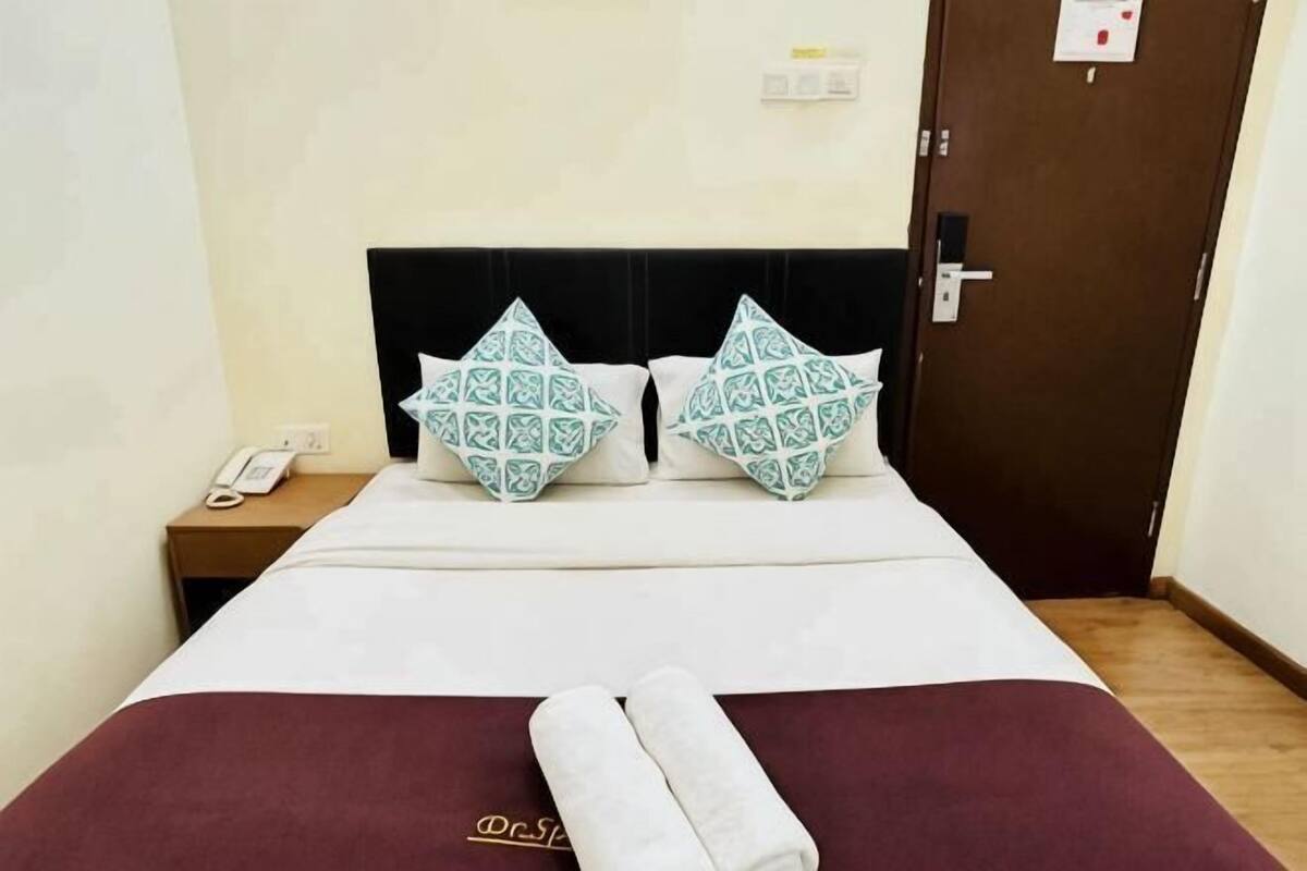 A hotel room with a bed, two patterned pillows, a neatly folded towel, a bedside phone, and a door.