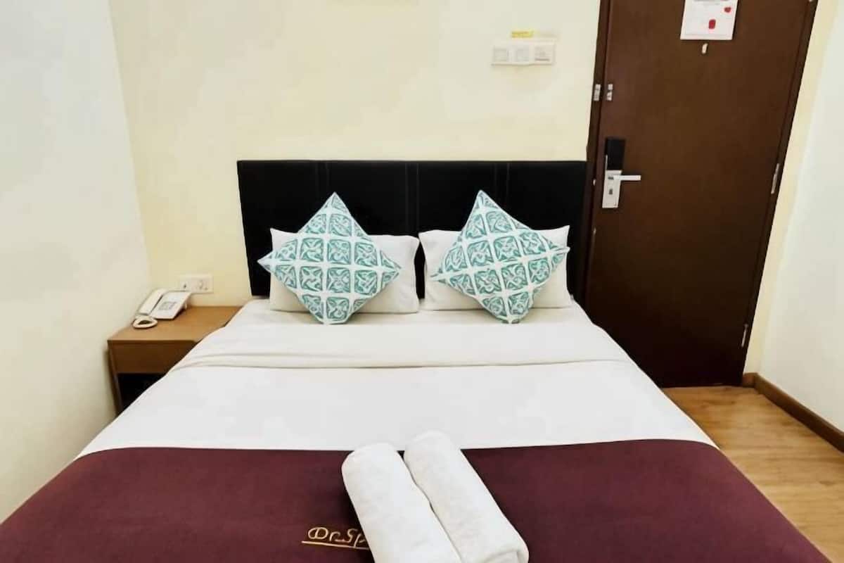 Quarto de hotel com uma cama, duas almofadas estampadas, uma toalha dobrada e um telefone ao lado da cama, além de uma porta.