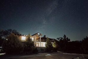 Une grande maison à deux étages, dotée d’un balcon, entourée d’arbres et baignée par la lumière d’un ciel étoilé.