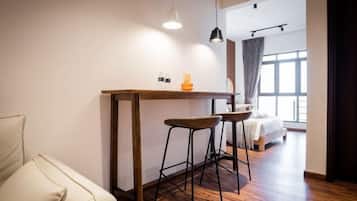 Quarto moderno com mesa de madeira, dois banquinhos, um sofá, uma cama e uma janela grande.