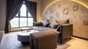 A modern living room with a sofa, ottoman, and a round coffee table. There is a large window with curtains, and a unique wall decoration.