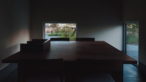 Ein schlichter Essbereich mit einem langen Tisch und Bänken, einem großen Fenster mit Blick auf Grünflächen und einem dunklen Boden.