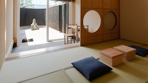 Ein Raum mit Tatami-Matten, verbunden mit einem offenen Bereich durch eine Schiebetür. Es gibt einen Holztisch, einen Stuhl und einen runden Spiegel an der Wand.