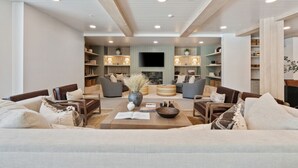 Uma sala de estar moderna com um sofá, poltronas, uma mesa de centro e livros. Há estantes com objetos decorativos e uma televisão de tela plana na parede.
