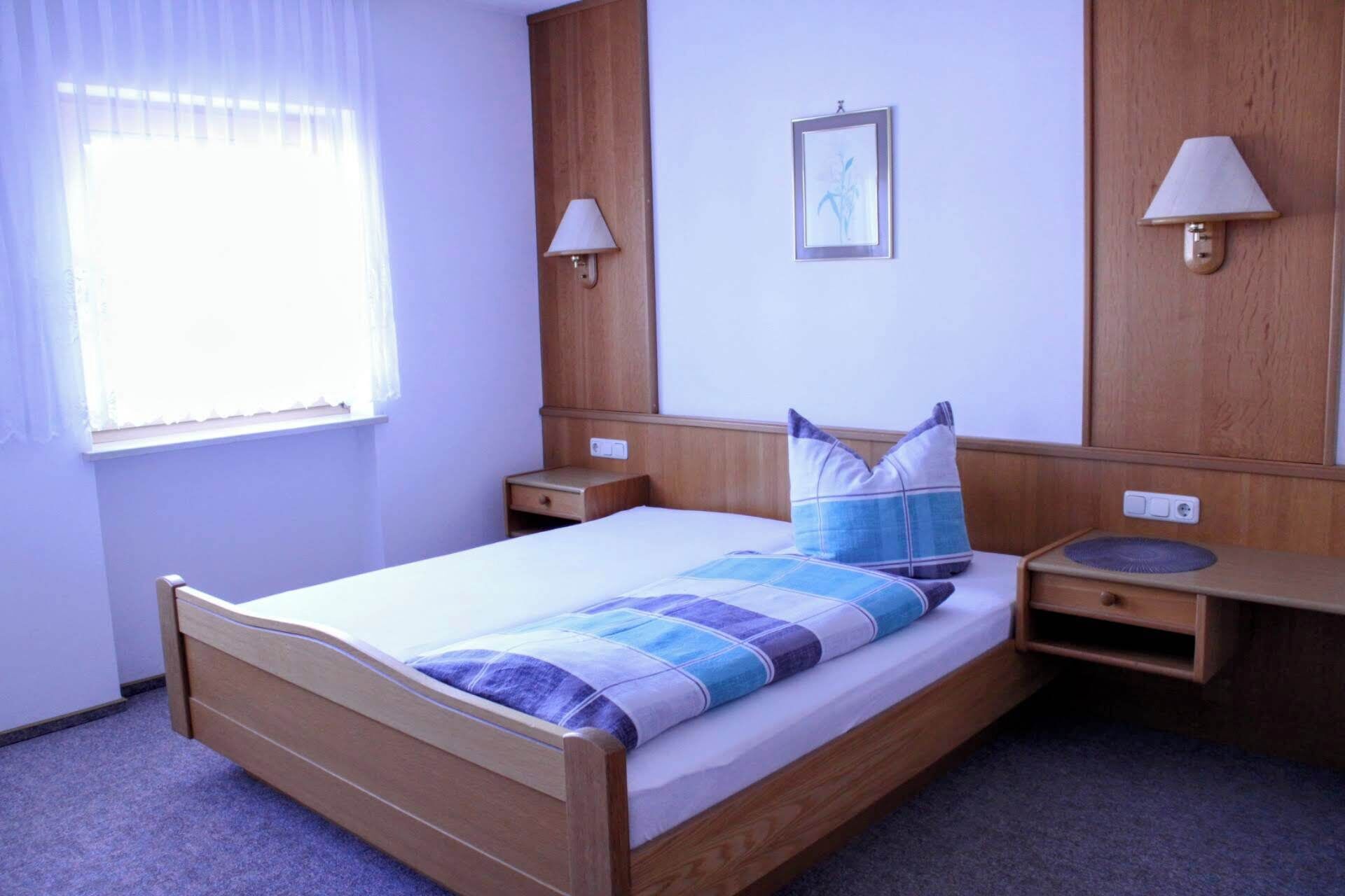 A single bed with a blue and white striped bedspread, two bedside tables, a nightstand, a window with sheer curtains, and two wall-mounted lamps.
