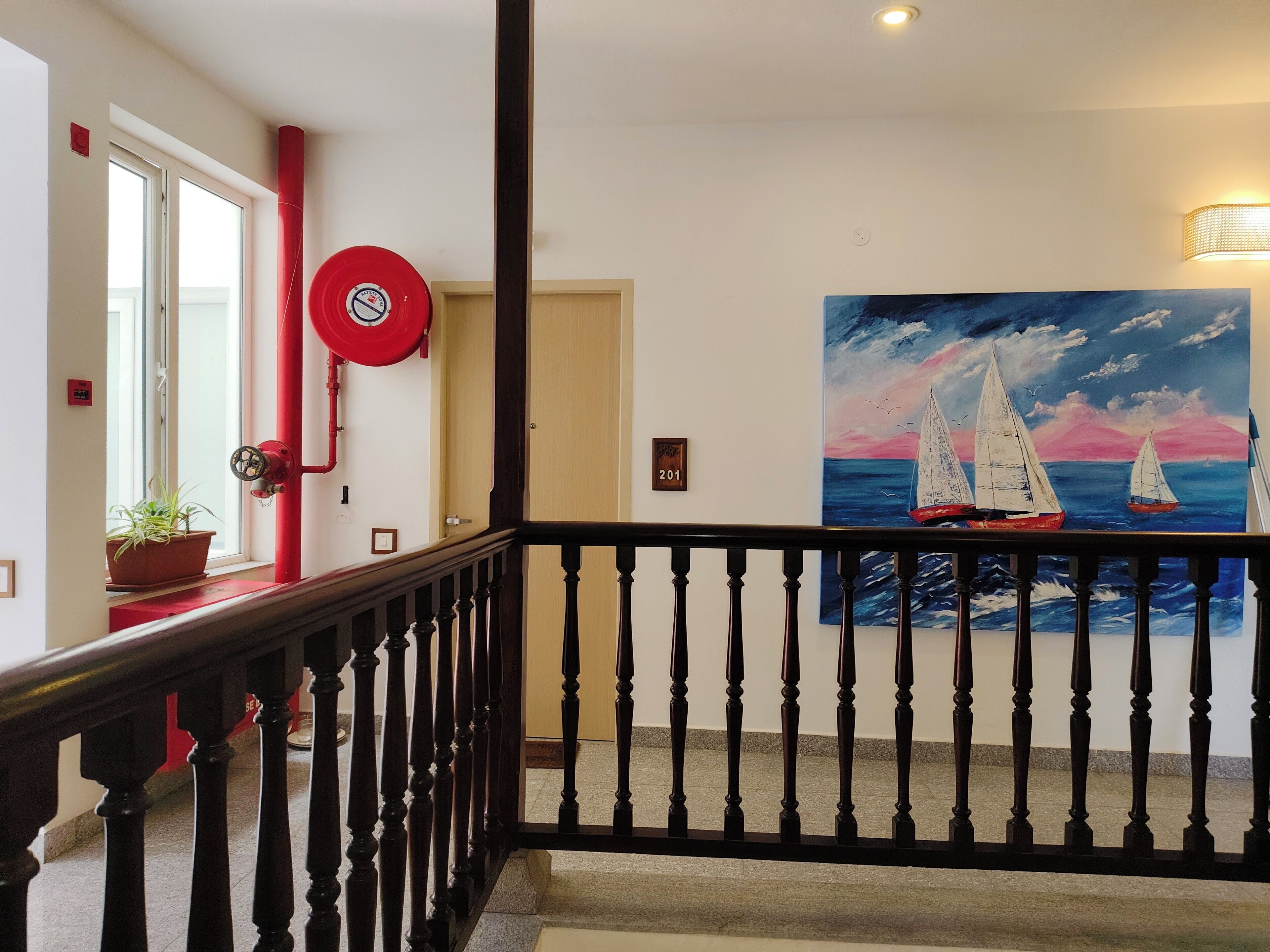 Un escalier avec une rampe, un tableau représentant des voiliers et un détecteur d’incendie rouge fixé au mur.