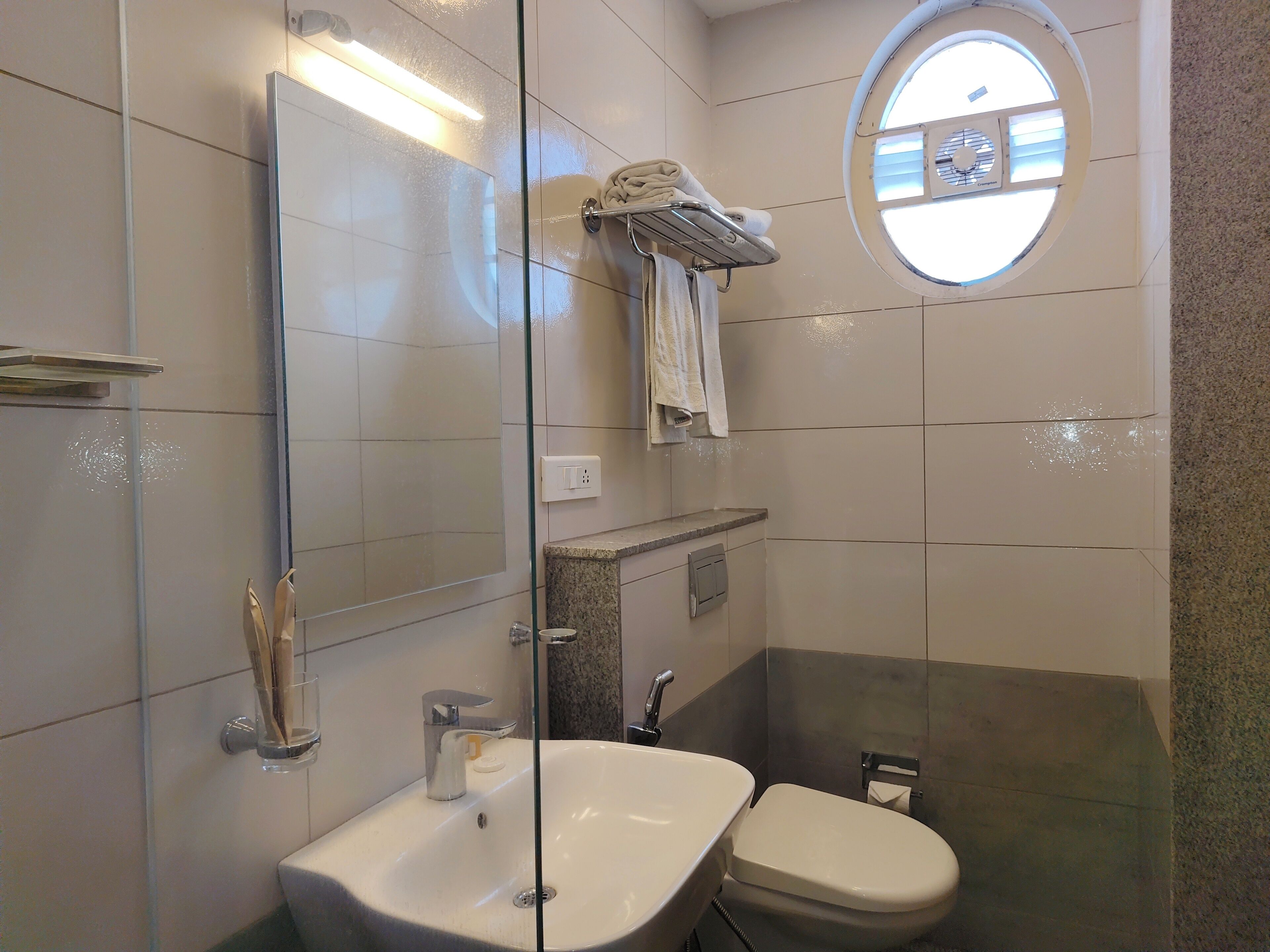 Un baño moderno con ducha envuelta en vidrio, lavamanos blanco y inodoro. Hay un perchero con toallas sobre el lavamanos y una ventana pequeña con vistas al cielo.
