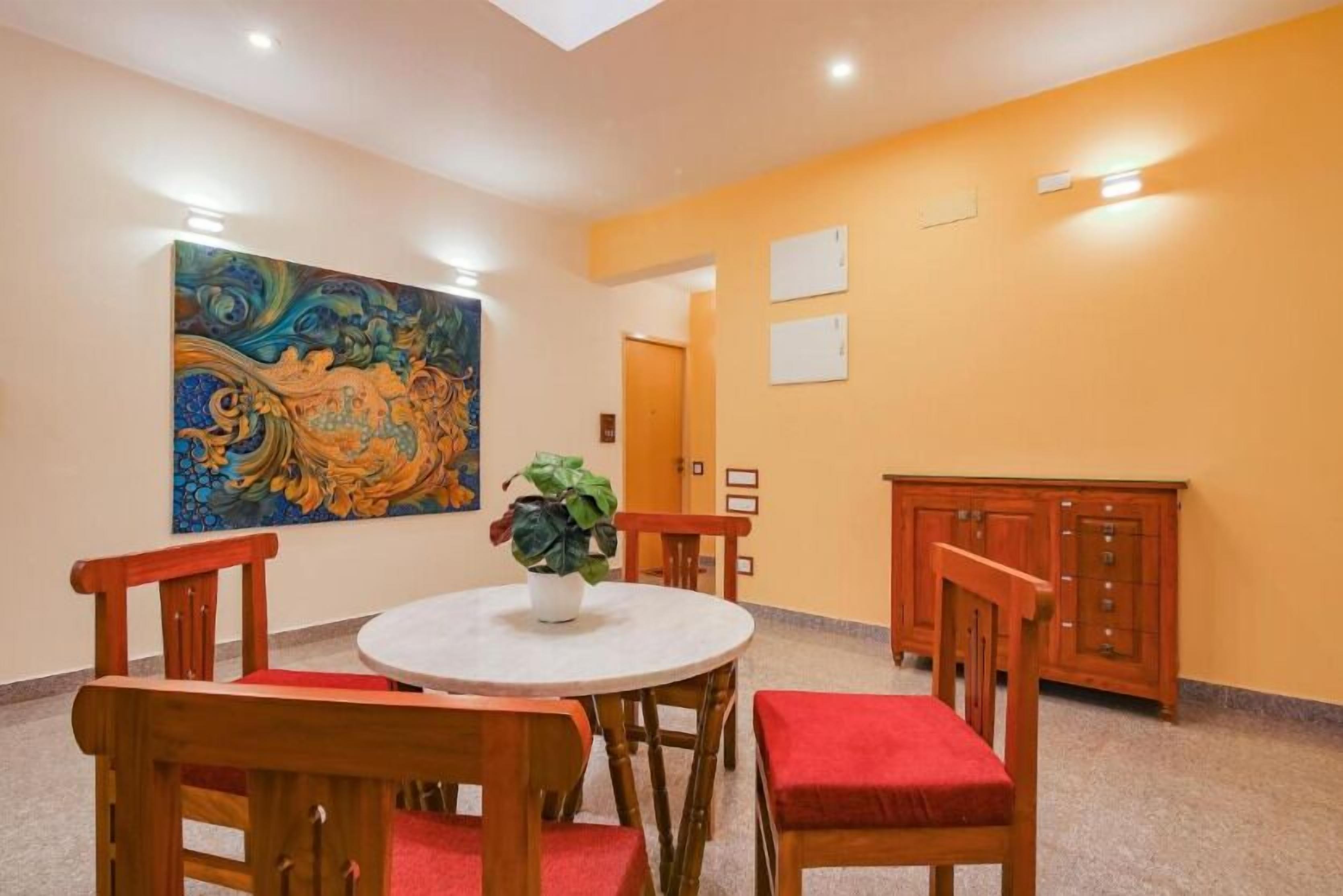 A dining area with a round table, wooden chairs, and a red cushioned bench. A large abstract painting adorns the wall, and a potted plant is on the table.