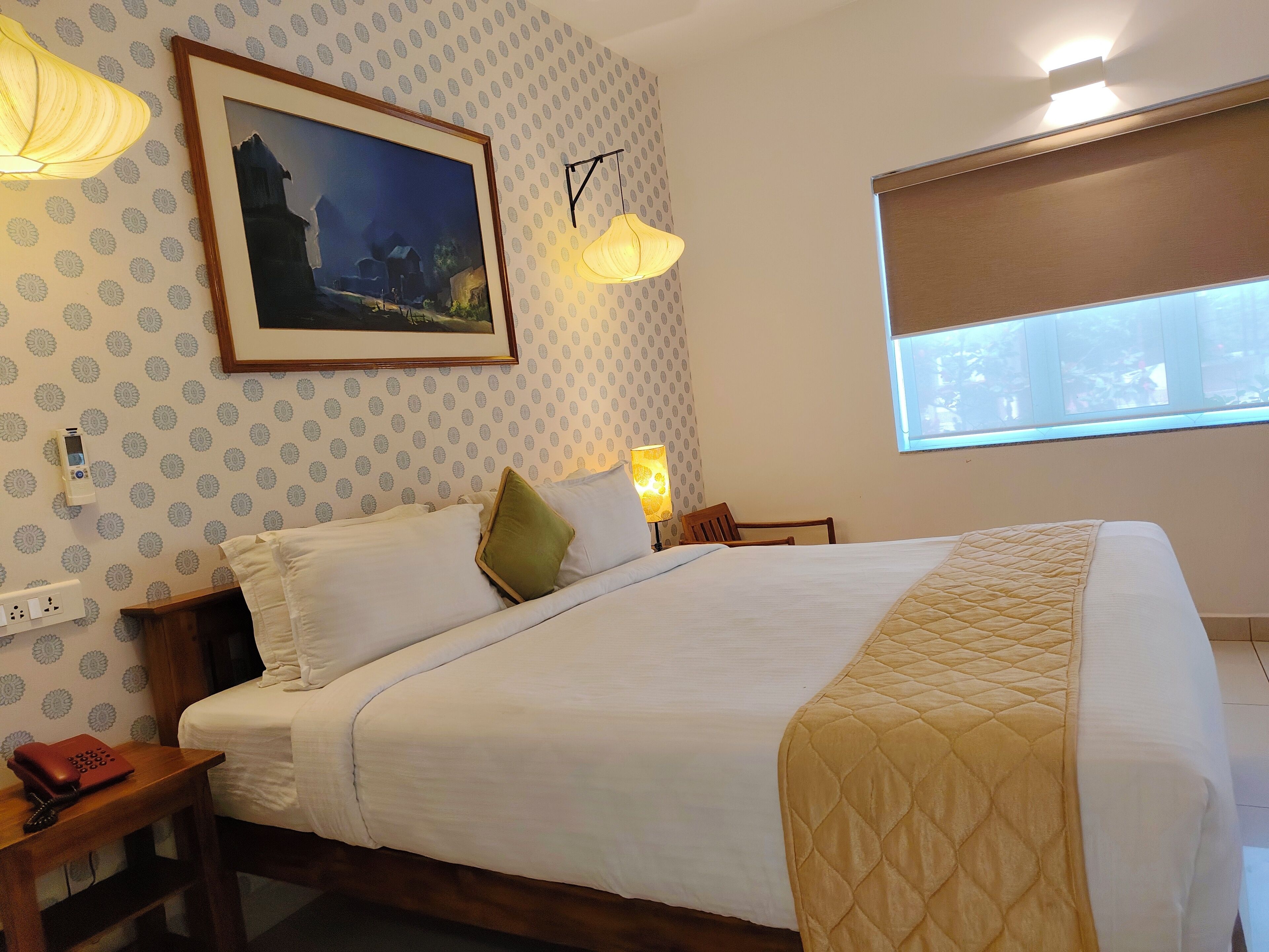 A neatly made bed with a quilted cover, a framed painting on the wall, a bedside lamp, and a window with blinds.
