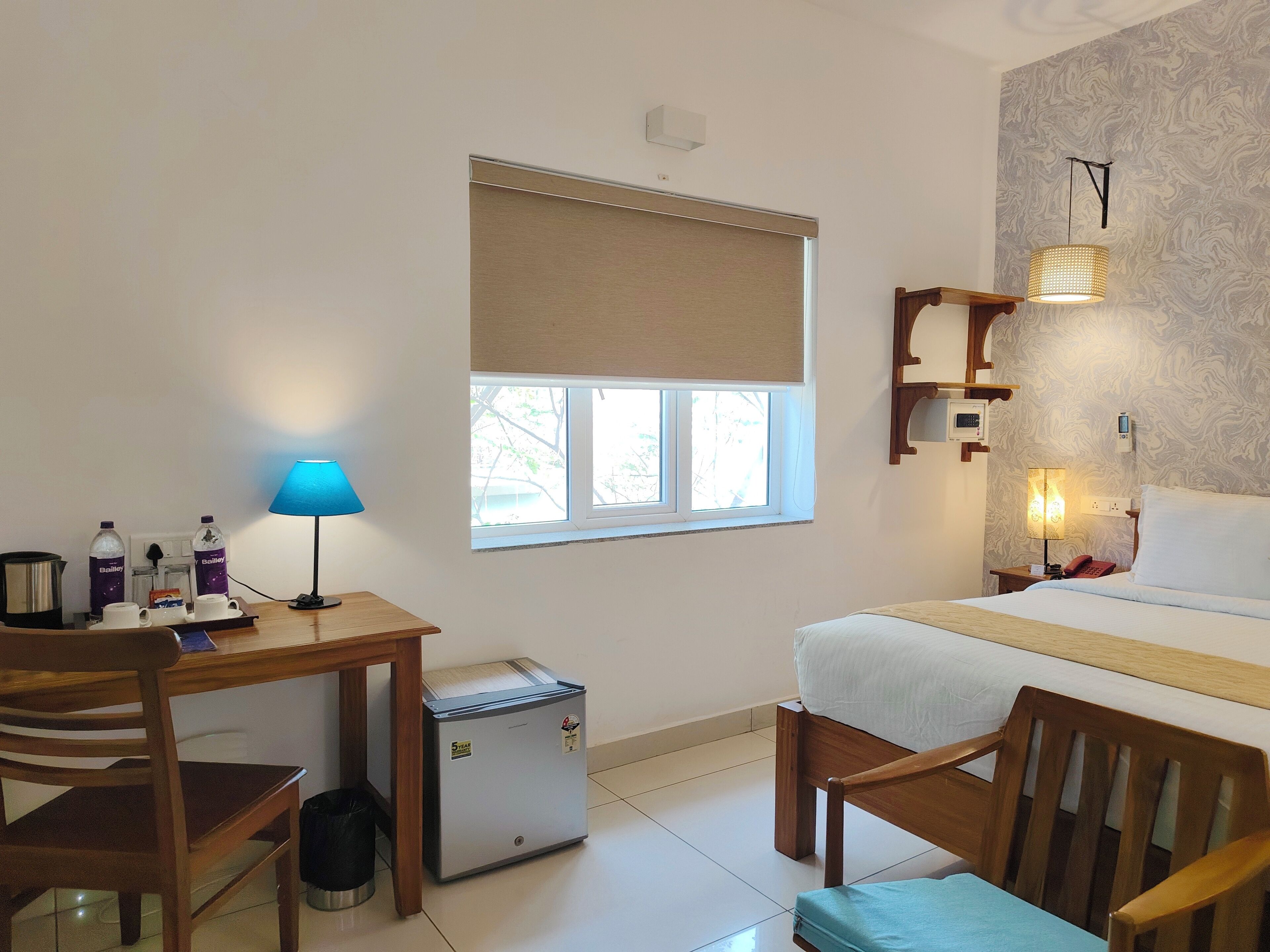 Une chambre d’hôtel avec un lit, un bureau doté d’une lampe, un petit réfrigérateur et une fenêtre donnant sur des arbres.