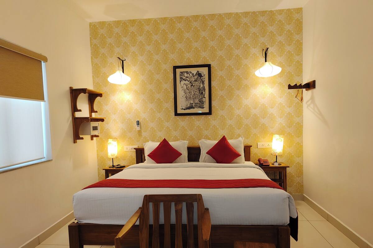 Una habitación de hotel con una cama grande, dos mesitas de noche, una silla de madera, una mesita pequeña y un cuadro colgado en la pared.