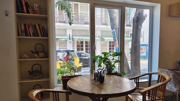 A cozy room with a round wooden table, two wooden chairs, a bookshelf with books, and a window with a view of the street and a tree outside. - Hotel Aura by Woco Stays (Puducherry)