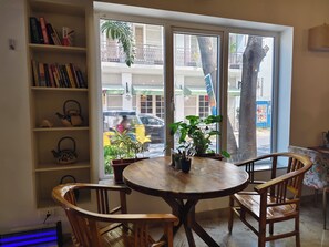 Une pièce chaleureuse avec une table ronde en bois, deux chaises en bois, une bibliothèque remplie de livres et une fenêtre donnant sur la rue et un arbre à l’extérieur.