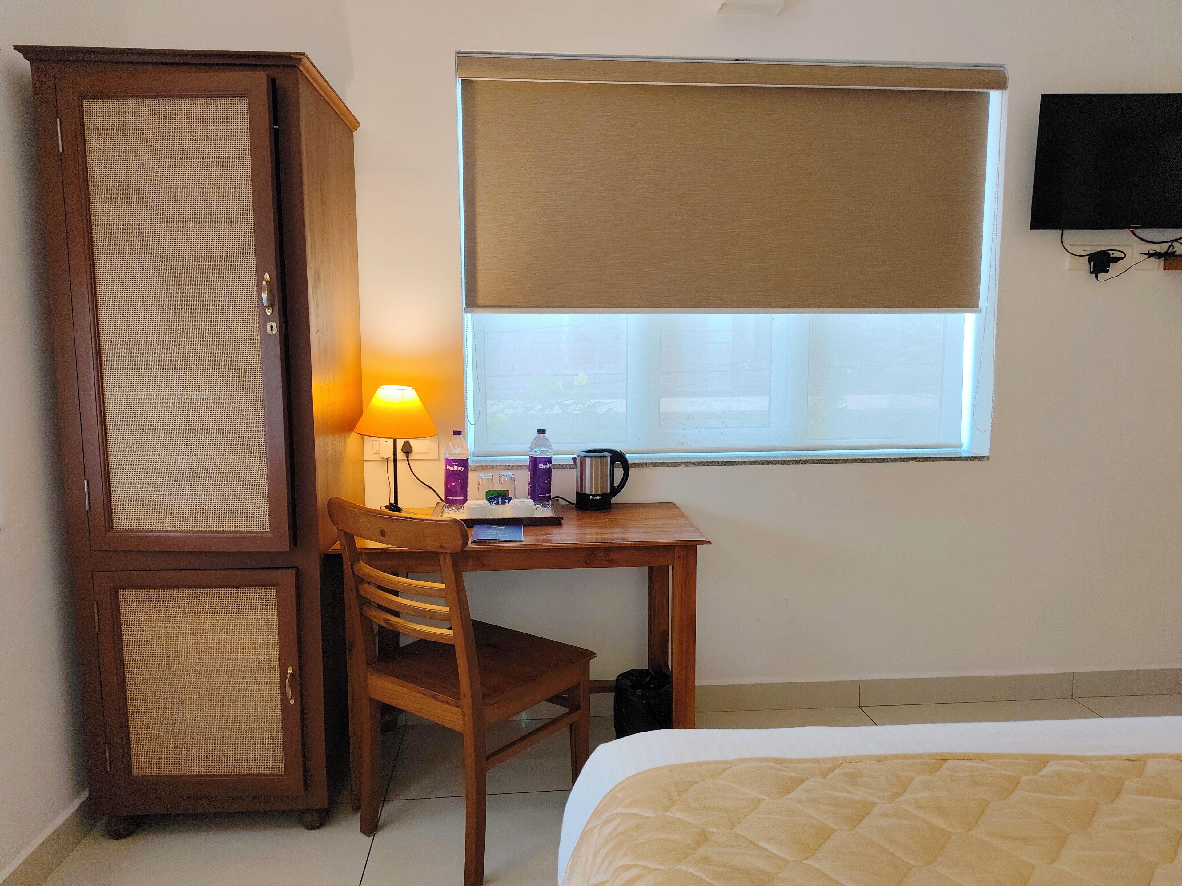 A hotel room with a bed, a wooden desk with a lamp, a chair, a wardrobe, and a window with a blind.