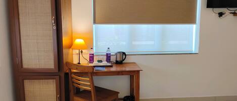 A hotel room with a bed, a wooden desk with a lamp, a chair, a wardrobe, and a window with a blind.