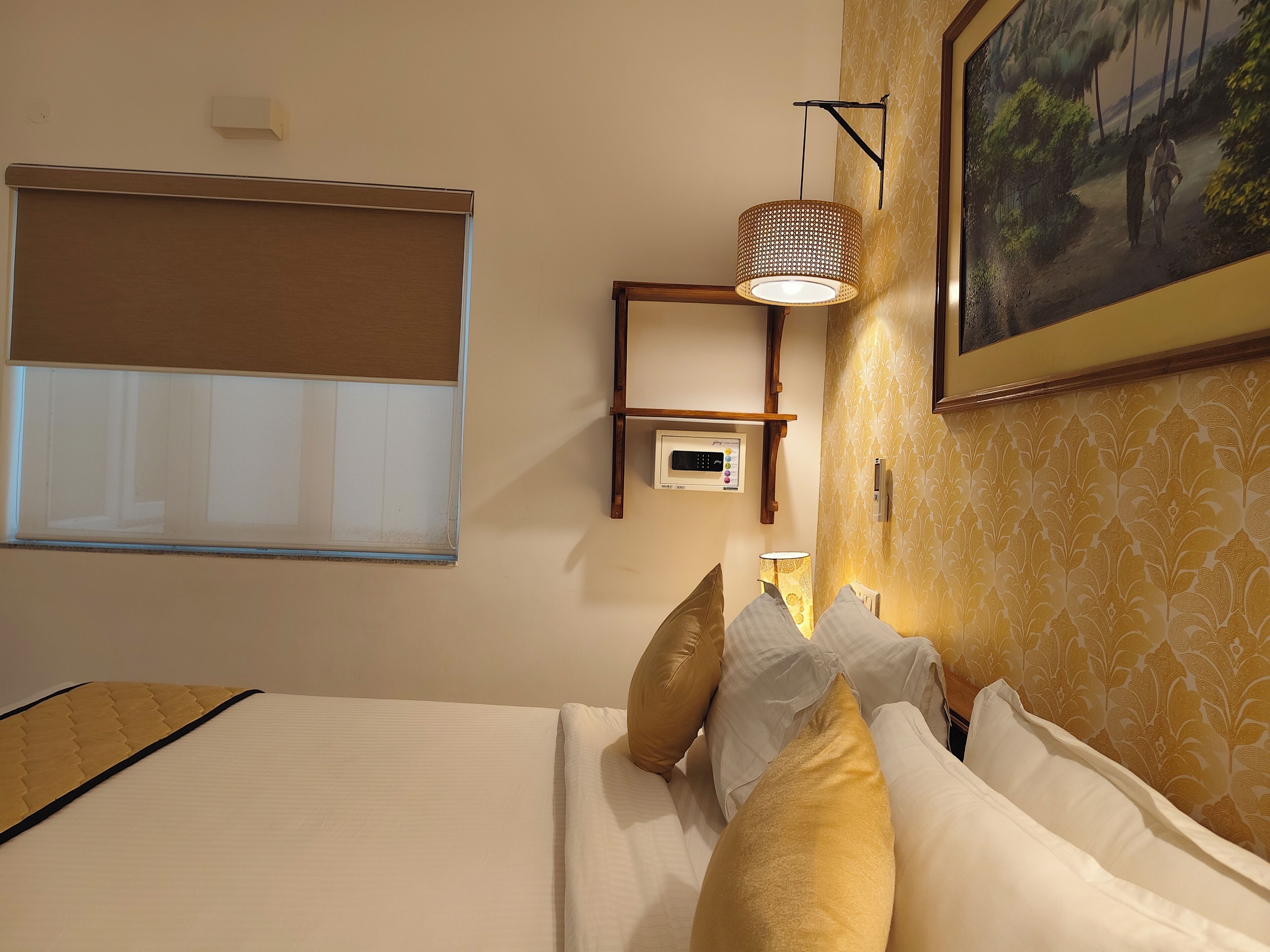 A hotel room with a bed, pillows, a wall-mounted lamp, a thermostat, and a window with a blind.